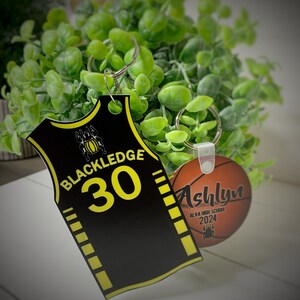 May include: Two keychains with basketball themes. One is a black jersey with the name "Blackledge" and the number "30" in yellow. The other is a basketball with the name "Ashlyn" and "ALVA HIGH SCHOOL 2024".