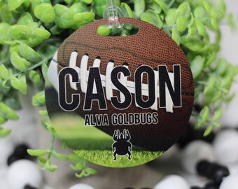 May include: Round luggage tag with a football design. The tag features the name "CASON" in black text and the words "ALVA GOLDBUGS" in smaller black text. A black and white bug illustration is at the bottom of the tag.