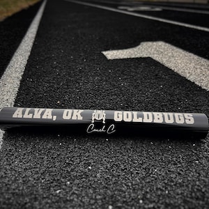 May include: A black track and field baton with the words "ALVA, OK GOLDBUGS" in silver lettering. The baton also has the words "Coach C" in a cursive font. The baton is lying on a black track with white lane markings.