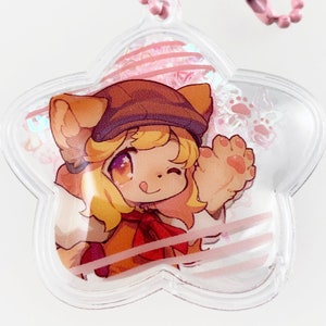 May include: A clear star-shaped keychain with a pink chain. Inside the keychain is a cartoon image of a character with blonde hair, wearing a brown hat and a red scarf. The character is waving with one paw.