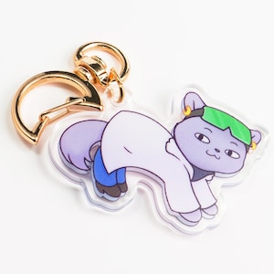 以下が含まれることがあります： 金色の金属製のリングが付いたキーホルダーに、緑色のヘッドバンドと白い白衣を着た紫色の猫が描かれています。