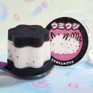 Op de afbeelding: Een zwart-wit dessertvormig object op een regenboog lepel. Een ronde roze en zwarte sticker met Japanse tekst en een cartoon illustratie van een zeeslak staat naast het dessert. Een doorzichtige plastic beker staat op de achtergrond.