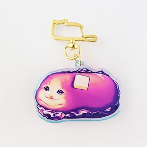 May include: A purple cat meme keychain with a gold metal clasp. The cat is depicted as a sandwich with a pat of butter on top.