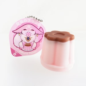 May include: A pink and brown pudding keychain with a charm featuring a pink fox character holding a cup of pudding. The charm has the text "狐のプリン sakura flavor" on it.