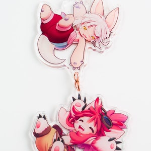 Può includere: Due ciondoli di animali dei cartoni animati rosa e bianchi su una catena dorata con un fermaglio a forma di cuore. I ciondoli sono di una creatura pelosa bianca con vestiti rossi e blu e una creatura pelosa rosa con vestiti rossi e gialli.