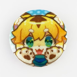 Pode incluir: Um botão redondo de pelúcia com um personagem de desenho animado com cabelo amarelo, olhos verdes e patas marrons. O personagem está vestindo uma camisa listrada azul e verde e está sorrindo.