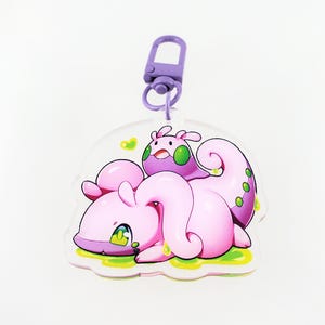 Peut inclure: Un porte-clés en acrylique transparent avec un personnage de dessin animé violet et rose et un cœur vert. Le porte-clés est doté d'un clip métallique.