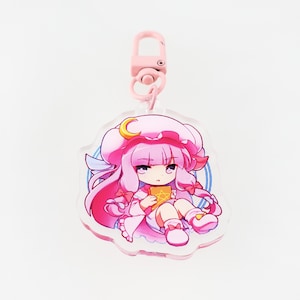 Könnte beinhalten: Ein rosa-blauer Anime-Charakter-Schlüsselanhänger mit einem Halbmond auf ihrem Hut. Sie hält ein Buch und hat eine rosa Schleife im Haar.