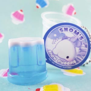 May include: A blue and white dessert cup with a matching tag that reads "Snom's Snow Pudding" with a cartoon character. The dessert is milk and coconut flavored. The background is a light blue with cupcake designs.