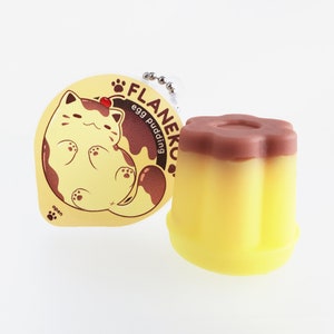 Può includere: Un gatto cartone animato giallo e marrone su un adesivo rotondo con il testo "FLANeko egg pudding open" e una piccola ciliegia rossa in cima.  Accanto all'adesivo c'è un budino giallo e marrone con una parte superiore a forma di fiore.