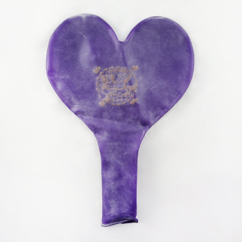May include: A purple heart-shaped balloon with a gold design in the center. The balloon is deflated and has a long, thin tail.