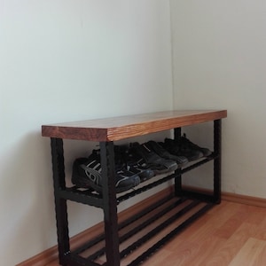 May include: A wooden and metal shoe rack with two shelves. The top shelf is made of light brown wood, and the frame is black metal. Several pairs of black shoes are on the shelves. The rack is placed against a white wall.