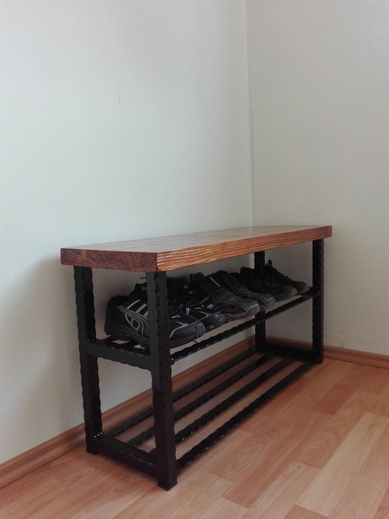 May include: A wooden and metal shoe rack with two shelves. The top shelf is made of light brown wood, and the frame is black metal. Several pairs of black shoes are on the shelves. The rack is placed against a white wall.
