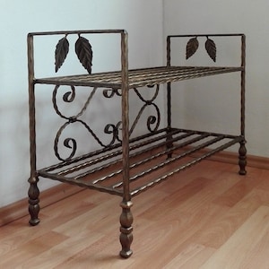 May include: A two-tiered wrought iron shoe rack with decorative leaf accents and scrollwork. The rack has a gold finish and stands on four legs.