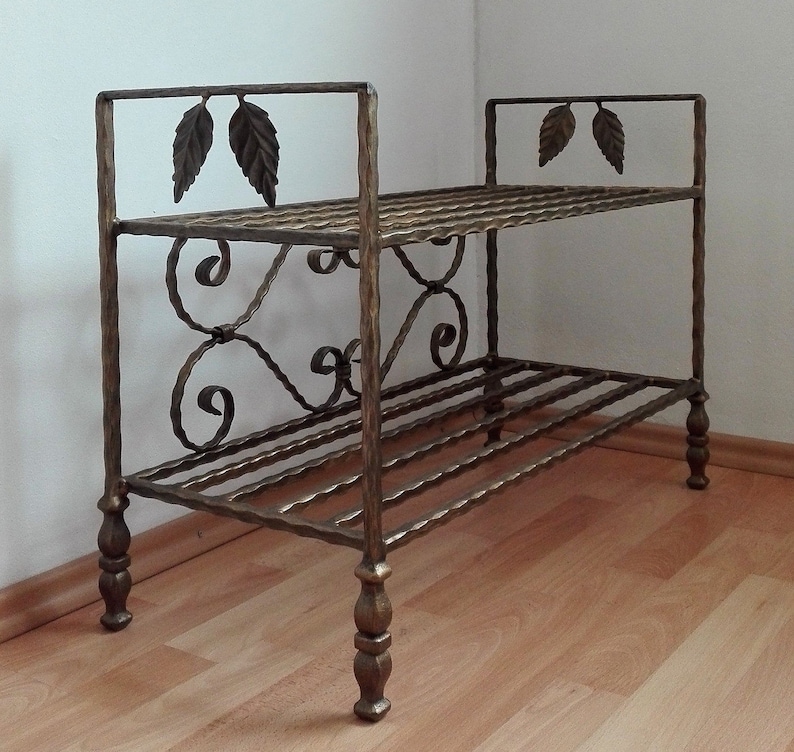 May include: A two-tiered wrought iron shoe rack with decorative leaf accents and scrollwork. The rack has a gold finish and stands on four legs.