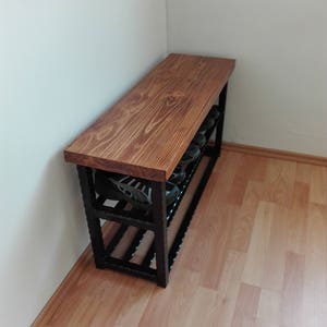 May include: A wooden shoe bench with a dark brown stained top and a black metal frame. The bench has two shelves for shoe storage and is placed against a white wall. The bench is approximately 48 inches long.