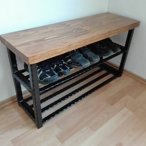 May include: A wooden shoe bench with a dark stained top and black metal frame. The bench has two shelves holding several pairs of shoes. The wood grain is visible on the top surface. The bench is placed against a white wall.