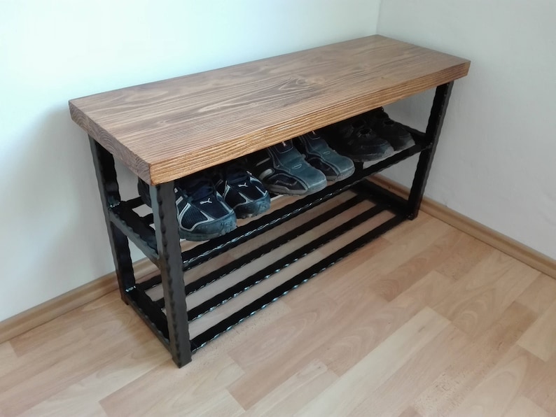 May include: A wooden shoe bench with a dark stained top and black metal frame. The bench has two shelves holding several pairs of shoes. The wood grain is visible on the top surface. The bench is placed against a white wall.