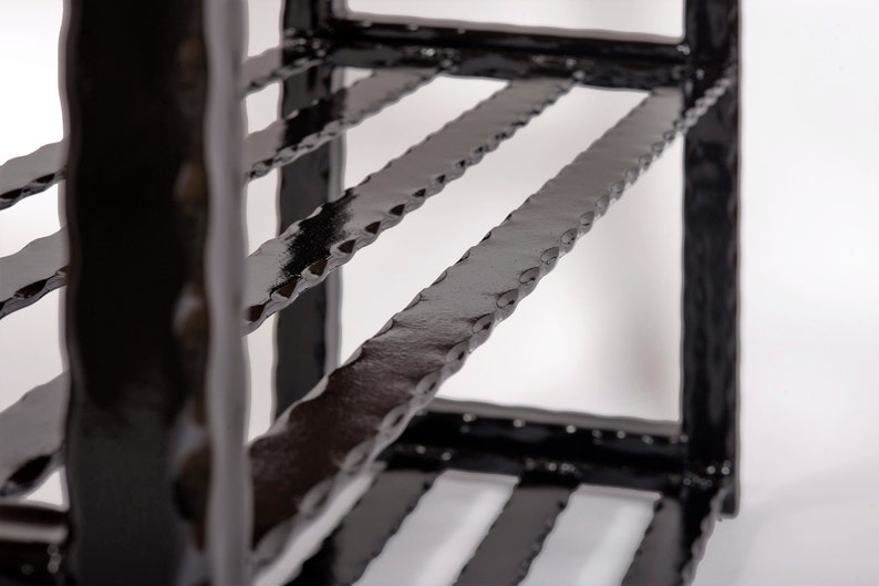 May include: Close-up of a black metal shelf with a textured, hammered finish. The shelf has multiple levels with horizontal bars and vertical supports. The design is industrial and modern, with a focus on the texture and the dark color.