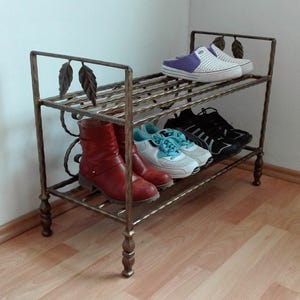 May include: A two-tiered metal shoe rack with a decorative leaf design. The rack is made of wrought iron and has a gold finish. The rack is filled with shoes, including boots, sneakers, and sandals.