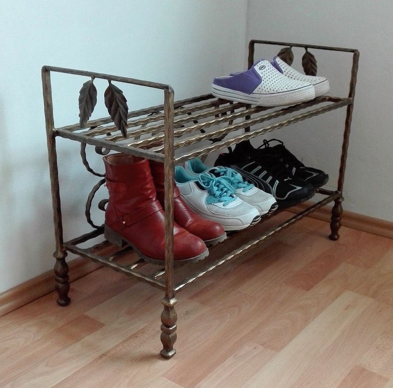 May include: A two-tiered metal shoe rack with a decorative leaf design. The rack is made of wrought iron and has a gold finish. The rack is filled with shoes, including boots, sneakers, and sandals.