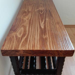 May include: A wooden bench with a stained, light brown top and a dark brown, decorative base. The bench top has visible wood grain and a textured surface. The base features vertical, dark brown supports.
