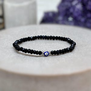 May include: A black beaded bracelet with a silver evil eye charm. The beads are small and round, and the charm is blue and white.
