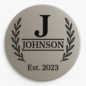 Może przedstawiać: Okrągła, szczotkowana metalowa tabliczka z czarnym napisem "JOHNSON", wyśrodkowanym pod dużą literą "J". Tekst "Est. 2023" znajduje się na dole. Wzory liści laurowych obramowują nazwę.