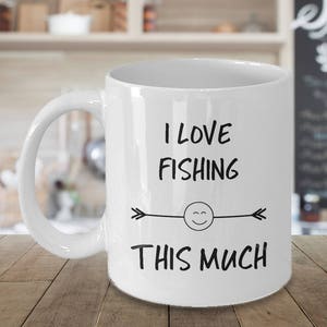 May include: White ceramic coffee mug with a black design that says "I love fishing this much" with a smiley face and arrows on either side.