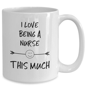 May include: White ceramic coffee mug with black text that reads "I love being a nurse this much" with a smiley face and arrows on either side.