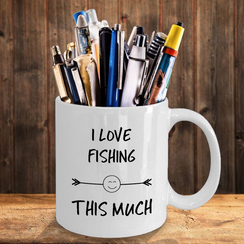 May include: White ceramic mug with a black design that says "I love fishing this much" with a smiley face and arrow. The mug is filled with pens and pencils.