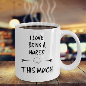 May include: White ceramic coffee mug with black text that reads "I love being a nurse this much" with a smiley face and arrows on either side of the smiley face.