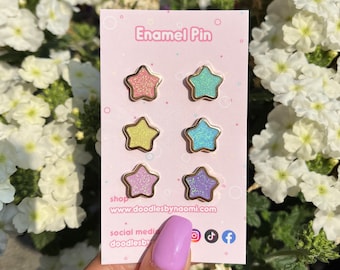May include: Set of six pastel glitter enamel pins shaped like stars with gold outlines. The pins are displayed on a pink card with the text "Enamel Pin" and the website address "www.doodlesbynaomi.com".