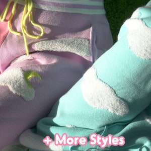 May include: Two pairs of sweatpants with fluffy cloud and moon appliques. One pair is pink with a yellow drawstring and the other is turquoise. The text " + More Styles" is visible at the bottom of the image.
