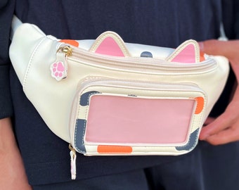 May include: A white fanny pack with a black, orange, and pink design. The pack has a clear plastic window on the front and a paw print charm.