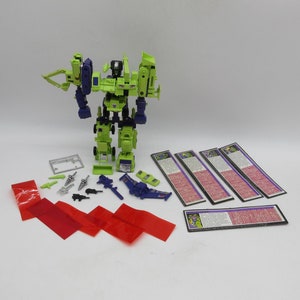 May include: A green and purple robot toy with various accessories, including weapons, a vehicle, and instruction cards. The robot is standing upright, with multiple articulated joints. Red plastic strips and small silver and black weapons are also included.