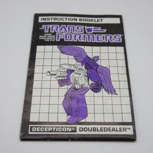 Könnte beinhalten: Anleitung für das Transformers-Spielzeug Doubledealer. Das Heft zeigt eine Schwarzweißillustration von Doubledealer im Robotermodus mit violetten Akzenten. Der Text auf dem Heft lautet "The Transformers" und "Decepticon Doubledealer".