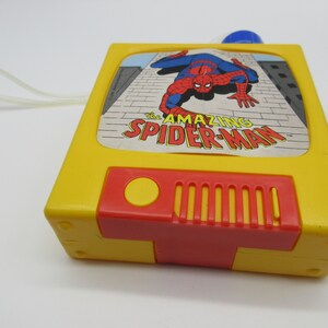 May include: A yellow plastic toy television with a red button and a speaker. The screen shows a cartoon image of Spider-Man in red and blue, with the text "The Amazing Spider-Man".