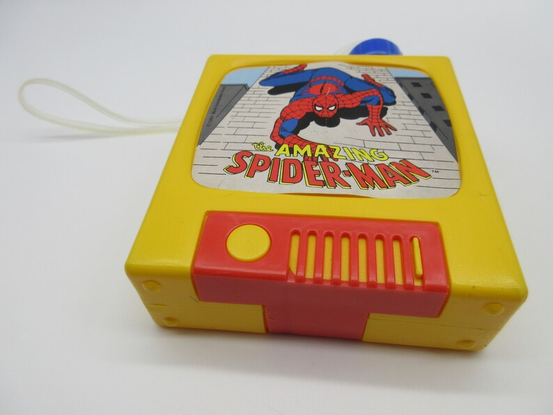 May include: A yellow plastic toy television with a red button and a speaker. The screen shows a cartoon image of Spider-Man in red and blue, with the text "The Amazing Spider-Man".