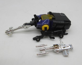 May include: A black, purple, and yellow toy robot with a silver weapon attached. Another silver weapon is lying next to the robot. The toy has wheels and a cockpit. The toy is on a white surface.