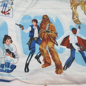 May include: A white fabric sheet featuring a Star Wars print with illustrations of Princess Leia, Han Solo, Chewbacca, and Lando Calrissian. The characters are depicted in action poses against a light blue background with additional mechanical details.