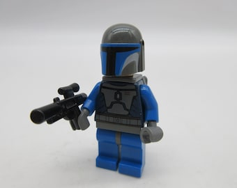 May include: A blue and gray Lego minifigure of a Mandalorian warrior with a black blaster rifle. The minifigure has a blue helmet with a gray visor and a gray chest plate with a blue stripe.