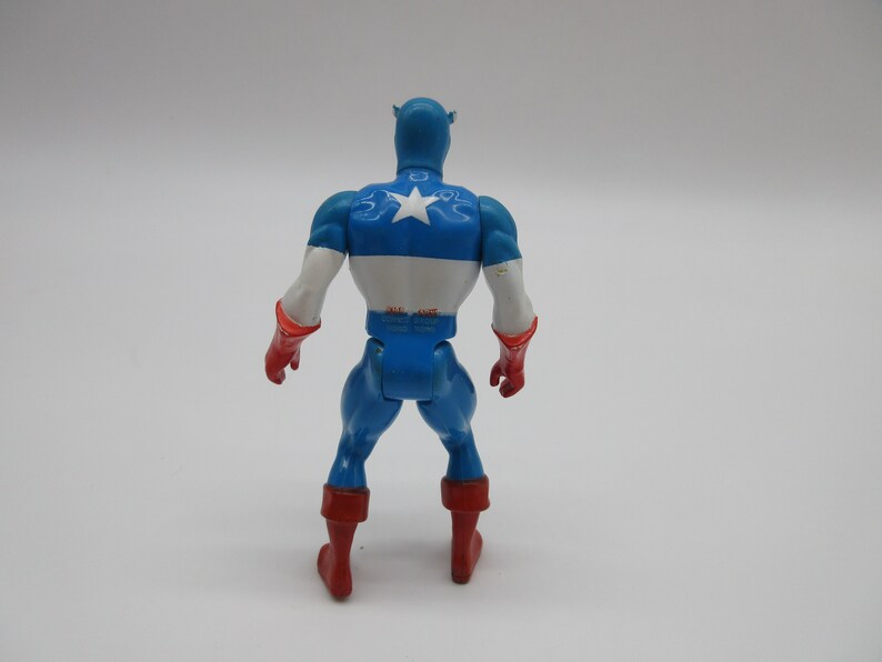 May include: A blue and red plastic action figure of Captain America. The figure is wearing a blue and white costume with a red star on the chest. The figure is standing with its arms at its sides.