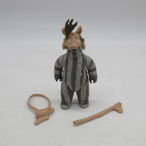 Op de afbeelding: Een grijs en wit gestreepte actiefiguur van een wezen met een varkensachtig masker en een groen, gewei-achtig hoofddeksel. Accessoires zijn een beige lus en een houten knots. De figuur staat op een witte ondergrond.