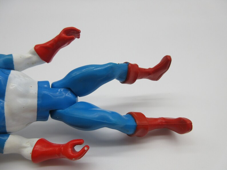 May include: A plastic action figure of a superhero wearing a blue and white costume with red boots and gloves. The figure is lying on its back with its legs spread apart.