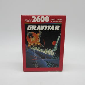 Puede incluir: Una caja de cartucho de videojuego Atari 2600 Gravitar vintage. La caja roja presenta el título del juego y un gráfico colorido de una nave espacial y un planeta. El logotipo de Atari es visible en la parte superior izquierda.