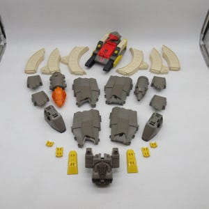 May include: A collection of plastic toy robot parts including a small tank vehicle, several curved beige track segments, various gray armor plates, two gray engine components with orange tips, and small yellow accent pieces arranged on a surface.