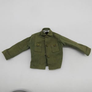 May include: An olive green miniature long-sleeve shirt with a collar, two front chest pockets, and three buttons down the center. The interior neck label features the text G.I. JOE in black lettering on a white background.