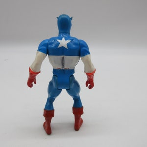 May include: A blue and red plastic action figure of Captain America. The figure is standing with its arms at its sides and is facing away from the camera. The figure has a white star on its chest.