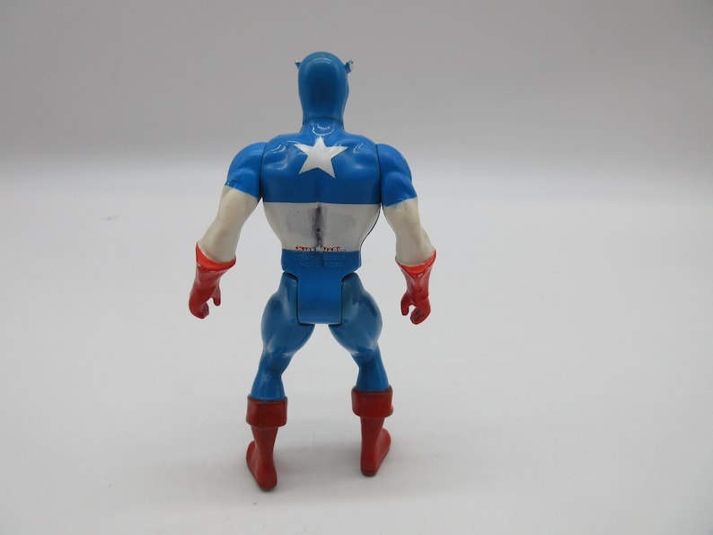May include: A blue and red plastic action figure of Captain America. The figure is standing with its arms at its sides and is facing away from the camera. The figure has a white star on its chest.